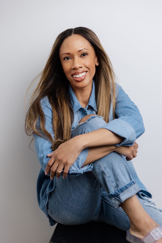 Woman in denim outfit sitting cross-legged, smiling warmly.